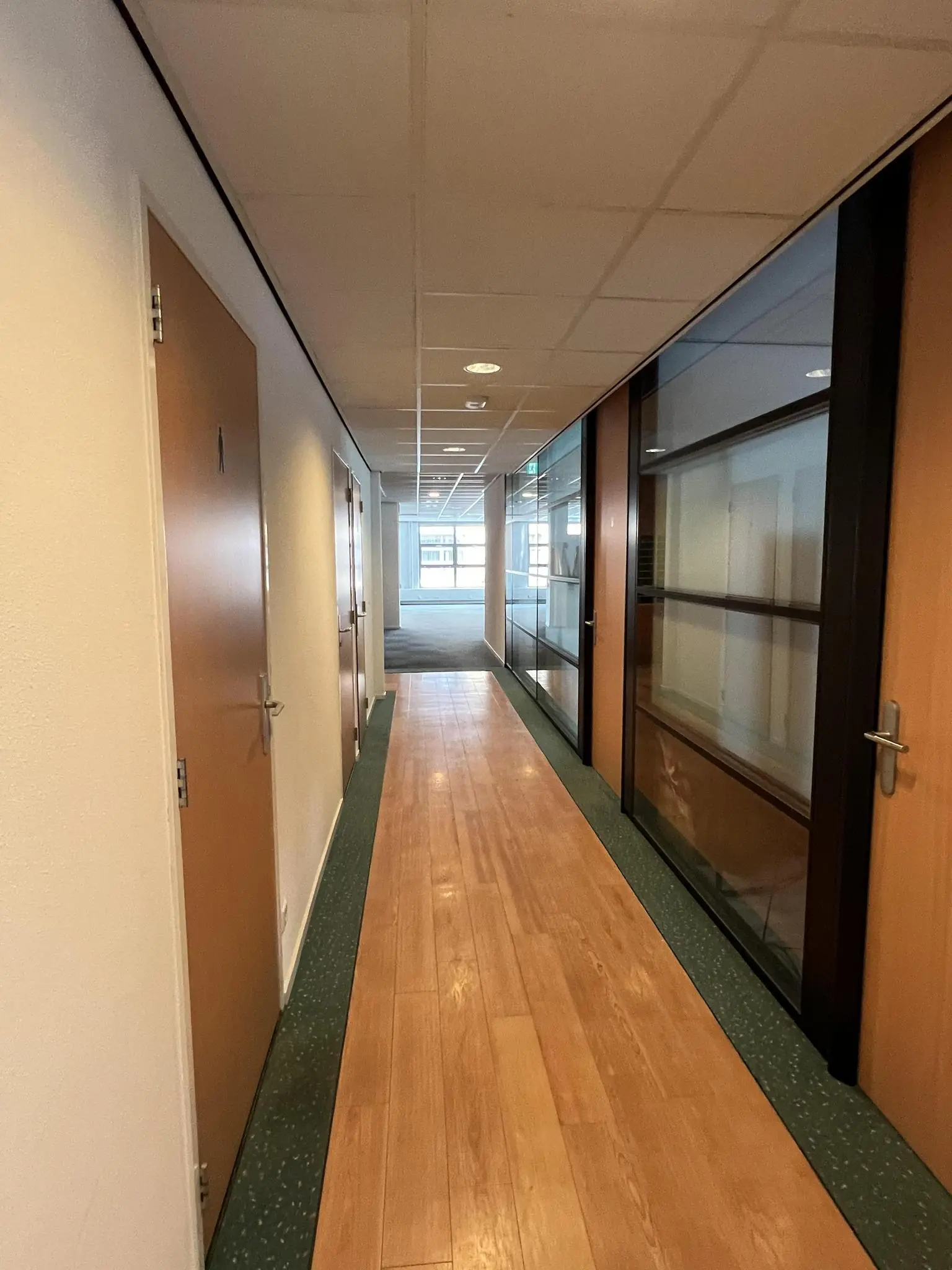 Gang in een kantoorpand aan de Rivium 2e Straat met houten vloer, deuren aan beide zijden en glazen kantoorwanden rechts.